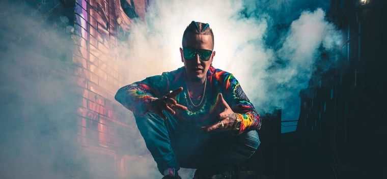 Sirreal crouching in drifting smoke in front of a brick wall, in a tie-dye jacket and green mirrored sunglasses, hands held out toward the camera