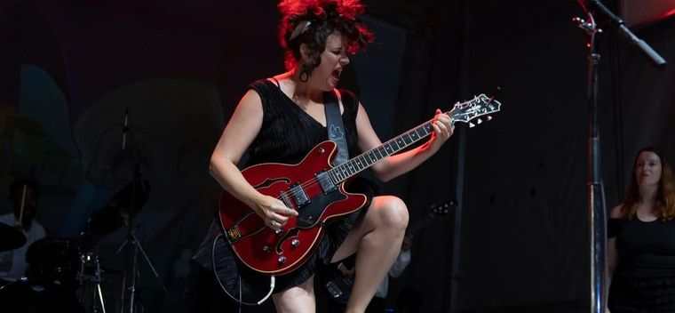 Kimberley MacGregor mid song on a dark stage, playing a red hollow body guitar with one knee raised