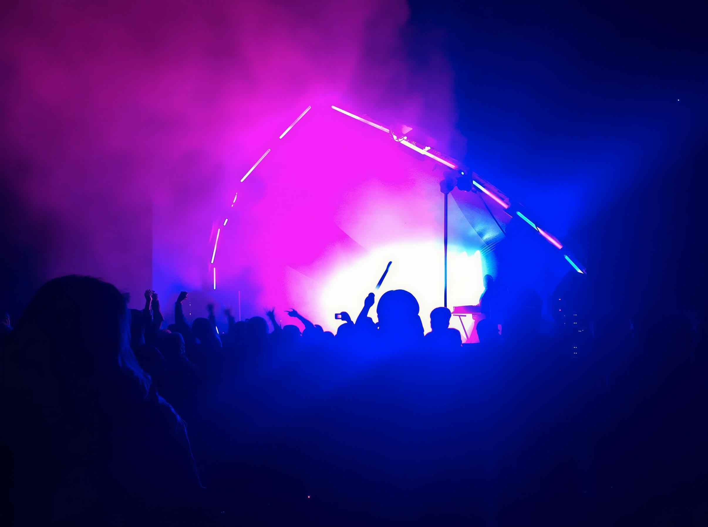 A crowd in silhouette, some with hands raised, in front of a stage glowing magenta and blue through smoke