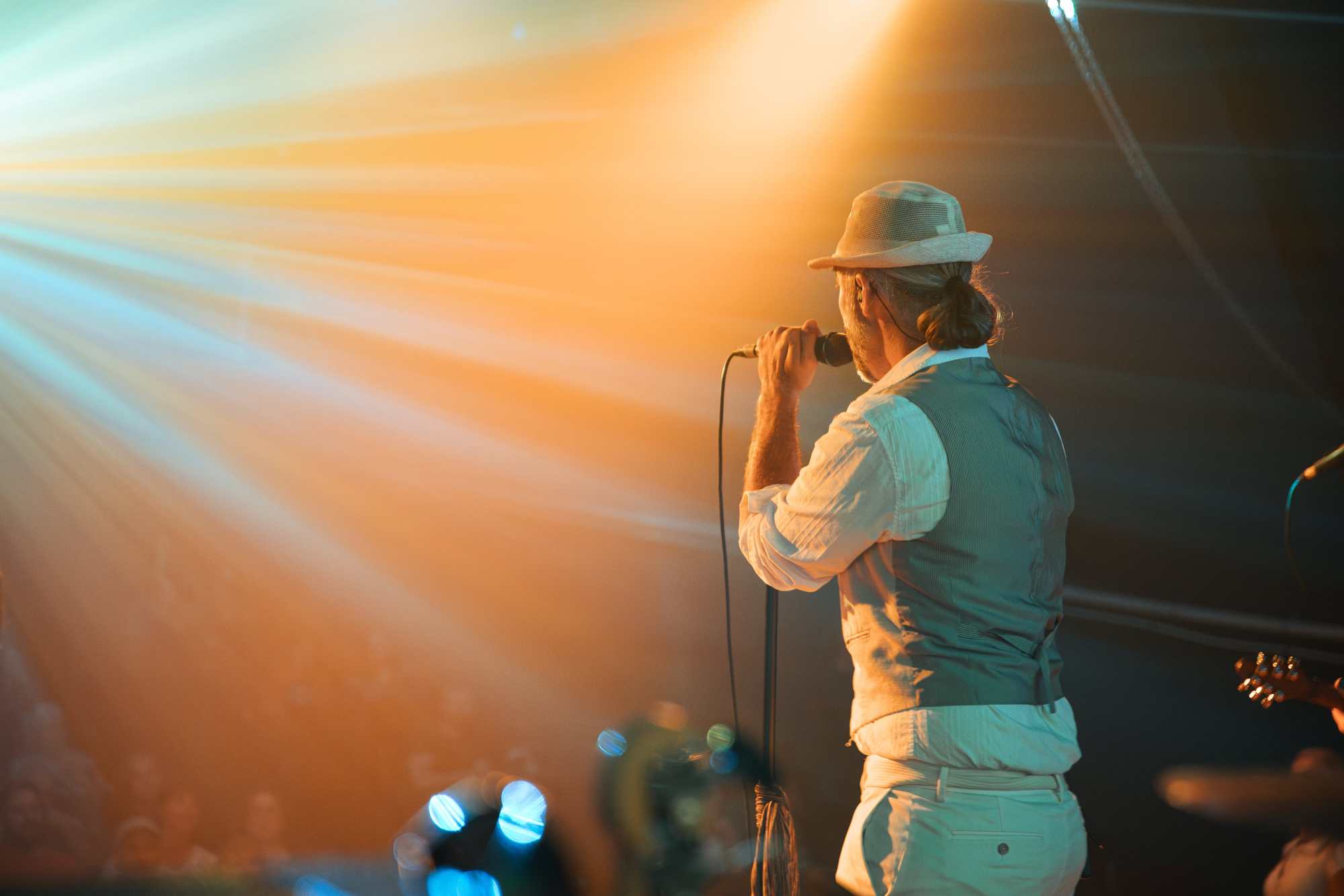 A singer in a hat and waistcoat, seen from behind, holding a microphone in a beam of warm orange stage light